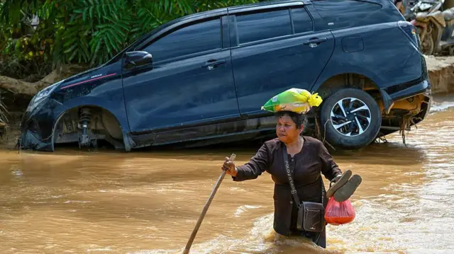 Seorang warga melintasi genangan banjir sambil membawa barang kebutuhan, sementara sebuah mobil terseret arus dan terperosok di pinggir jalan pascabanjir besar yang melanda wilayah Sumatra.